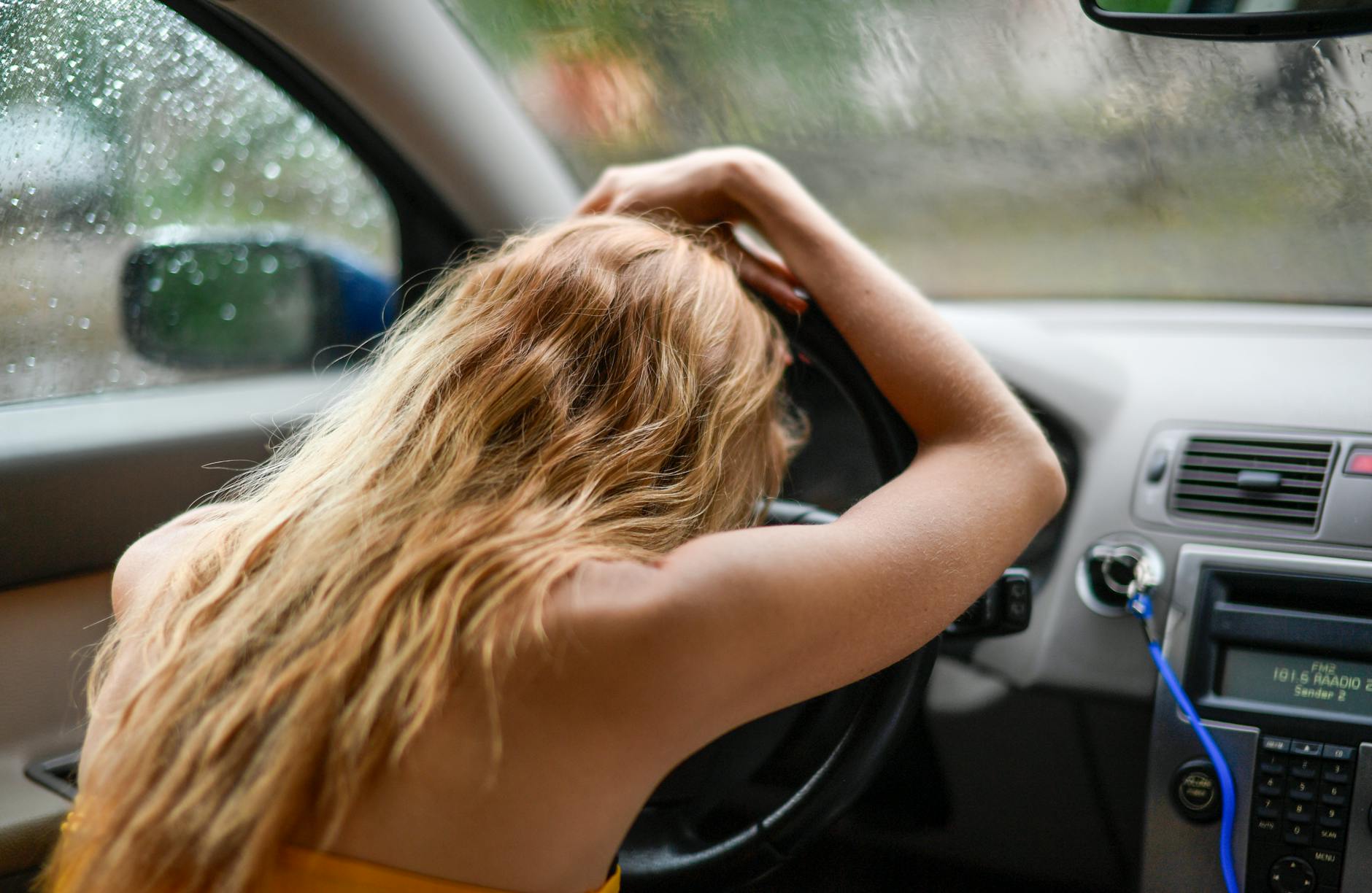 Zamyślona kobieta z blond włosami opiera głowę o kierownicę w aucie